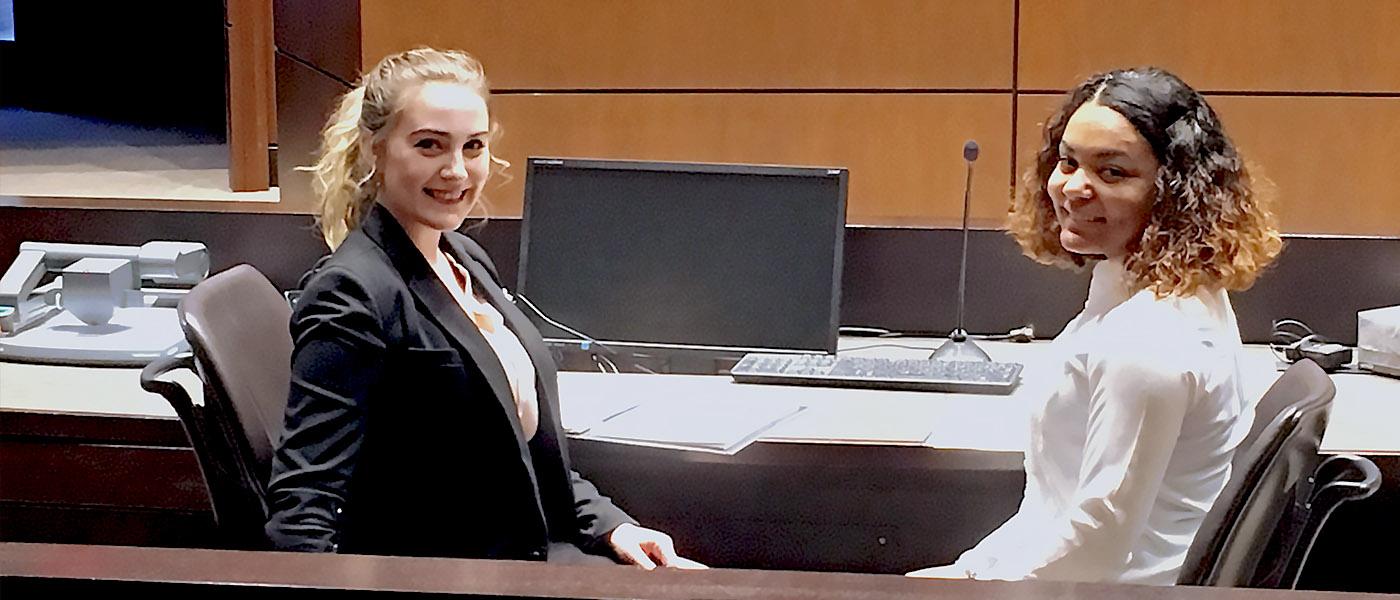 两名学生坐在法庭的法律顾问桌前，对着镜头微笑